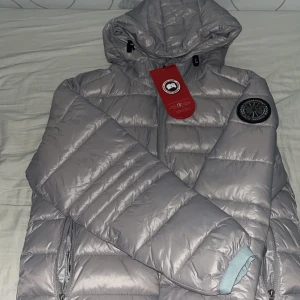Silver pufferjacka från Canada Goose - Säljer en snygg silverfärgad pufferjacka från Canada Goose med huva och svart patch på ärmen. Aldrig använd, helt ny. Pris kan diskuteras!