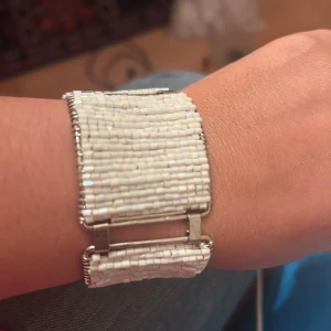 Brett vitt pärlarmband med silverdetaljer - Unikt och brett armband med små vita pärlor i flera rader, fäst på silverfärgade metallramar. Armbandet har en cool, geometrisk design och stängs med en metallkedja. Perfekt för dig som gillar statement-smycken och vill sticka ut med något annorlunda.