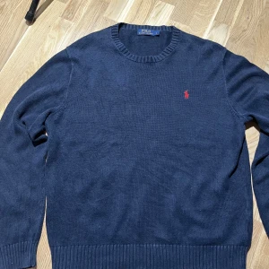Marinblå stickad tröja Polo Ralph Lauren - Snygg marinblå stickad tröja från Polo Ralph Lauren i storlek L. Klassisk rund hals och ribbade muddar vid ärmslut och nederkant. Tröjan har det ikoniska röda Polo-logot broderat på bröstet. Perfekt för dig som gillar stilrena och tidlösa plagg.