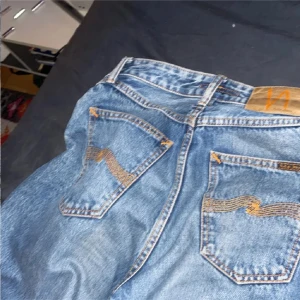 Blå Nudie Jeans W26 L30 - Säljer ett par klassiska blå jeans från Nudie Jeans med snygga kontrastsömmar och ikonisk broderad bakficka. Jeansen är i 100% bomull och har en normal passform. Perfekta för dig som gillar stilrena och tidlösa jeans med schyssta detaljer.
