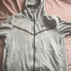 Grå hoodie från Nike med dragkedja - Grå hoodie från Nike i storlek L med hel dragkedja och huva. Tröjan har ett diskret mönster med en tunn linje över bröstet och Nike-logga på vänster sida. Tillverkad i mjuk bomull och polyester, perfekt för chill dagar. Långärmad och med fickor på sidorna.den har tre små skador, en skada syns lite mer men annars i väldigt bra skick