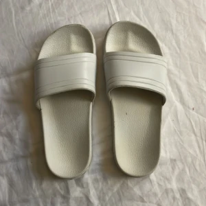 Vita slides sandaler - Säljer ett par stilrena vita slides sandaler med bred rem över foten och platt sula. Perfekta för chill dagar och enkla att glida i. Materialet är syntet och designen är minimalistisk med diskreta linjer på remmen.