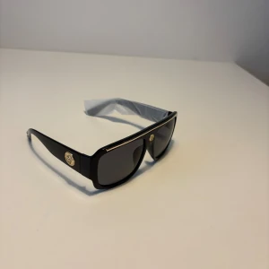 Versace liknande solglasögon  - Snygga svarta solglasögon från Versace med bred båge och rektangulär form. Glasögonen har mörka linser och coola guldfärgade Medusa-emblem på sidorna samt en tunn guldfärgad detalj längs ovansidan. Perfekt accessoar för en trendig look. Helt nya.