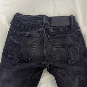 Tiger of Sweden svarta jeans Slender 25/32 - Svarta jeans från Tiger of Sweden i modellen Slender. Snygg tvätt med slitningar och klassiska fem fickor. Jeansen har smal passform och normal midja, med detaljer som silverfärgade nitar och svart läderpatch bak i midjan.