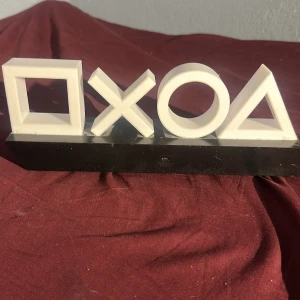 PlayStation Symbol Dekoration - Snygg PlayStation-dekoration med de klassiska symbolerna: fyrkant, kryss, cirkel och triangel. Perfekt för spelrummet eller som samlarpryl. Produkten är i gott skick med mindre damm och ytliga repor på basen. Officiell PlayStation-logga på foten.