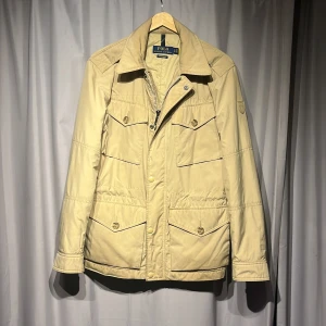 Beige vindjacka från Polo Ralph Lauren - Snygg beige vindjacka från Polo Ralph Lauren med fyra framfickor med lock och knappar, klassisk krage och logotyp på ärmen. Jackan har dragkedja och knappstängning framtill, samt justerbara ärmslut. Jackan kostar 3000kr nypris från Ralph lauren. Jackan sitter som storlek S.