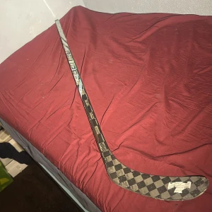 Bauer Tracer P28 40 Flex Hockeyklubba - Bauer Tracer P28 hockeyklubba med 40 flex. Kolfiberblad med schackmönster, lätt och slitstark. Viss kosmetisk slitage syns på skaftet, men klubban är i gott skick och redo för spel. Perfekt för juniorer eller spelare som söker en lättare flex.