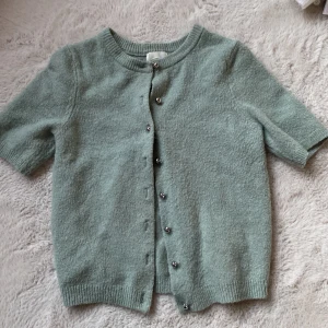 Mintgrön kofta från H&M XS - Superfin mintgrön kofta från H&M i storlek XS. Koftan har korta ärmar, rund hals och snygga silvriga knappar framtill. Materialet är mjukt och stickat, perfekt för lager-på-lager. Passformen är normal och koftan har ribbade muddar vid ärmslut och nederkant.