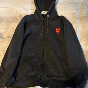 Svart hoodie från Comme des Garçons Play - Väldigt fet Svart hoodie från Comme des Garçons Play med dragkedja och huva. På bröstet finns två röda broderade hjärtan med ögon, en ikonisk detalj från märket. Tröjan har snörning vid huvan och två fickor framtill. Perfekt för dig som gillar streetwear och snygga detaljer.