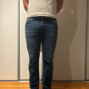 Mörkblå jeans från Jack & Jones - Snygga mörkblå jeans från Jack & Jones med klassisk femficksdesign och knappgylf. Jeansen har en straight passform och subtila slitningar framtill för en cool look. Perfekta för dig som gillar stilrena och bekväma jeans.