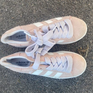 Adidas Campus rosa sneakers mocka - Adidas Campus sneakers i ljusrosa mocka med vita detaljer och klassiska tre ränder på sidan. Skorna har tjocka vita snören, platt sula och rund tå. Adidas-logga på hälen och insidan. Perfekt för dig som gillar retrostil och mjuka färger.