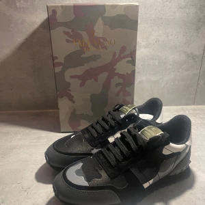 Valentino Garavani Rockrunner sneakers - Snygga Valentino Garavani Rockrunner sneakers med svart, grå och vit camouflage-mönster. Skorna har rund tå, platt sula och coola nitar på hälen. Materialmix av skinn och syntet med snörning och grönt logomärke på plösen. Perfekt för dig som gillar streetstyle och exklusiva märken.