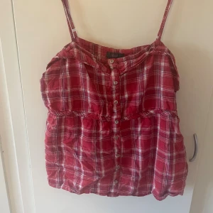 Rutig röd linnetopp från Dce - Säljer en röd och vit rutig linnetopp från Dce i storlek 46. Toppen har smala axelband, knappar framtill och en avslappnad passform. Perfekt för varma dagar när du vill ha en chill och snygg look.