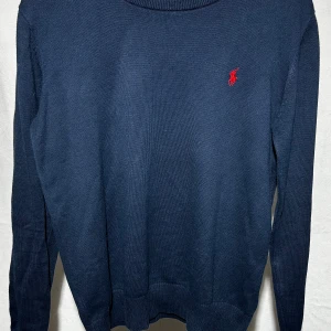 Marinblå stickad tröja från Polo Ralph Lauren - Snygg marinblå stickad tröja från Polo Ralph Lauren med klassisk röd logga broderad på bröstet. Tröjan har rund hals och långa ärmar, perfekt för dig som gillar stilrena och tidlösa plagg.