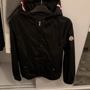 Svart vindjacka från Moncler - Svart vindjacka från Moncler med huva och dragkedja framtill. Jackan har Moncler-logga på ärmen och snygga rödvita detaljer på huvan. Tillverkad i lätt polyester, perfekt för blåsiga dagar. Klassisk passform och stilren look.