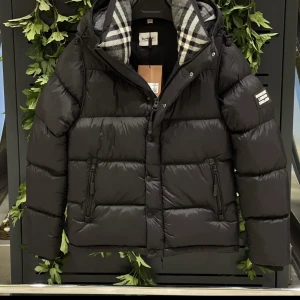 Svart pufferjacka från Burberry - Svart pufferjacka från Burberry med avtagbara ärmar, så du kan bära den som väst. Jackan har dragkedja, knappar och två fickor med zip. Insidan har klassiskt rutigt Burberry-foder i grått och vitt. Perfekt för kalla dagar med stil.