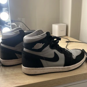 Nike Air Jordan 1 High svart/grå - Nike Air Jordan 1 mif sneakers i svart och grått med klassisk Air Jordan-logga på sidan. Skorna har snörning, rund tå och platt sula. Materialmix av skinn och mocka med pälsdetaljer på panelerna. Snygg kontrast mellan färgerna och ikonisk Nike Air-tunga.