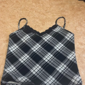 Svart och vit rutig linnetopp med spets - Säljer en snygg linnetopp med svart och vit rutig design. Toppen har tunna axelband och en v-ringad hals med svart spetskant. Den från USA så lite osäker vilken butik ifrån då jag köpt den för länge sen 