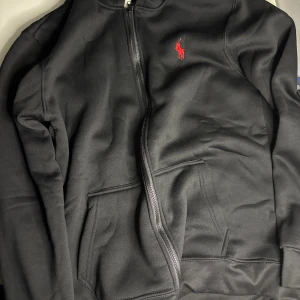 Svart hoodie från Polo Ralph Lauren - Svart hoodie från Polo Ralph Lauren med röd broderad logga på bröstet. Tröjan har dragkedja framtill, två fickor och huva med snörning. Materialet är mjukt och känns skönt mot huden, perfekt för chill dagar. Klassisk och stilren design.