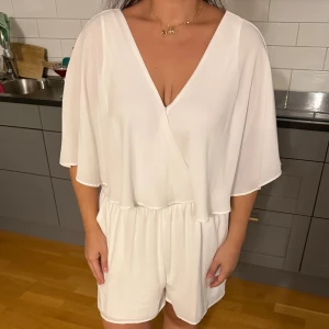 Vit jumpsuit  med snörning - Säljer en snygg jumpsuit med omlott-design och v-ringad fram. Shortsen har elastisk midja och är gjorda i ett mjukt, flowy material. Baksidan har öppen rygg med knytband för extra detalj. Perfekt för sommarens alla tillfällen.