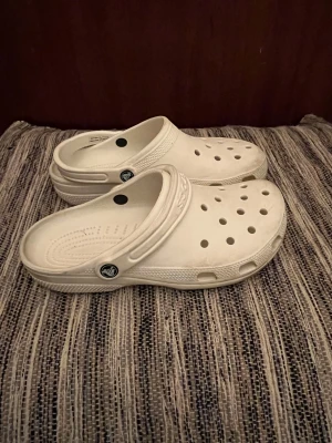 Vita Crocs tofflor med hålmönster - Säljer ett par klassiska vita Crocs sandaler med ventilerande hålmönster och justerbar hälrem. Storlek: M10/W8 = typ 42. Skorna är tillverkade i lätt syntetmaterial och har en rund tå samt platt sula. Perfekta för chill dagar och enkel att dra på sig.
