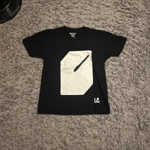 Svart t-shirt från Illuminated Apparel - Cool svart t-shirt från Illuminated Apparel med en stor vit panel på framsidan där du kan rita med medföljande penna som lyser i mörkret. T-shirten har rund hals och korta ärmar, tillverkad i 100% bomull. Perfekt för dig som gillar att vara kreativ och sticka ut. Passar till barn som är 9-11 år