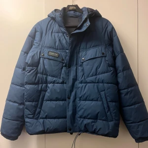 Blå Barbour pufferjacka med huva - Säljer denna riktigt feta vinterjackan från Barbour då den är lite stor. Jackan har huva, två stora bröstfickor med knappar, sidofickor och grov dragkedja. Materialet är syntet och jackan är quiltad för extra värme. Perfekt för kalla dagar och har en clean, modern look. Ovanlig färg och modell som knappt går att få tag på.