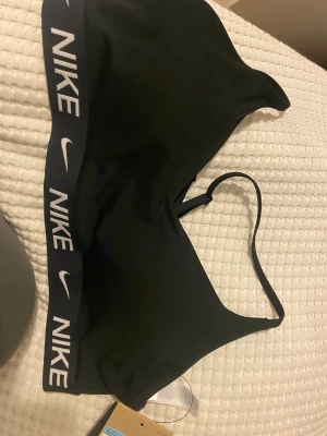 Svart Nike linnetopp med logoband - Nike Indy Bra i svart, med justerbara smala axelband och ett brett elastiskt band med vit Nike-logga runt om. Snygg och sportig design, perfekt för träning. Materialet är mjukt och stretchigt syntet för maximal komfort.