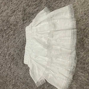 Vit volangkjol med spetsdetaljer - Supersöt vit kortkjol med flera lager volanger och spetsdetaljer längs kanterna. Kjolen har en elastisk midja som gör den bekväm och lätt att bära. Perfekt för dig som gillar romantisk och feminin stil.