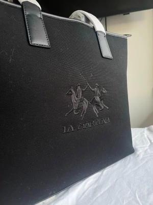 Svart handväska från La Martina - Helt ny svart handväska från La Martina med broderad logga och polospelare på framsidan. Väskan har svarta detaljer i skinn och robusta handtag. Materialet är slitstark bomull med inslag av skinn. Perfekt för dig som gillar klassisk design med sportig touch.