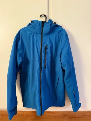 Five Seasons Skidjacka - Blå skidjacka med huva från Five Seasons, perfekt för dig som gillar att vara ute. Jackan har svart dragkedja framtill och på bröstfickan, samt flera praktiska innerfickor. Materialet är polyester och jackan har långärmad design med justerbara ärmslut.