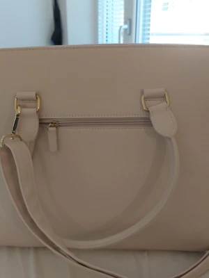 Beige axelväska med gulddetaljer - Snygg beige shopping/hand/axelväska med stilrena guldfärgade metalldetaljer och praktisk dragkedja både på utsidan och insidan. Väskan har flera fack och en avtagbar axelrem. Perfekt storlek för att få plats med allt du behöver under dagen.