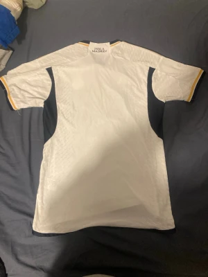 Real Madrid adidas matchtröja vit - Säljer en officiell Real Madrid matchtröja från adidas i vit med marinblå och gula detaljer. Tröjan har klubbmärke, adidas-logga och 'Emirates Fly Better' tryckt på bröstet. Tillverkad i polyester med HEAT.RDY-teknologi för extra komfort.