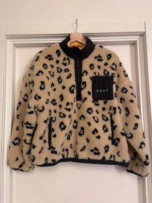OBEY Leopardmönstrad teddytröja - Beige teddytröja från OBEY med svart leopardmönster. Jackan har gult innefoder, svart krage och detaljer, dragkedja framtill och en svart bröstficka med OBEY-logga och dragkedja. Perfekt för dig som gillar streetwear och vill vara varm och samtidigt sticka ut med snygg stil. Originalpris: 1800kr. Mycket bra skick! Använd 2 ggr. Sälj på flera sidor, först till kvarn. 