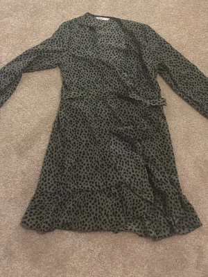 Leopardmönstrad omlottklänning från ONLY - Säljer en snygg leopardmönstrad omlottklänning från ONLY i storlek 34. Klänningen är svart och grå med långa ärmar och volangdetaljer längs nederkanten. Perfekt för dig som gillar trendiga mönster och vill sticka ut lite extra.