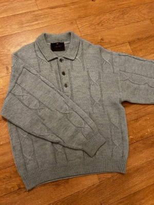 Grå stickad pikétröja med kabelmönster - Snygg grå stickad pikétröja från Pierre Balmain med klassiskt kabelmönster. Tröjan har krage, tre knappar framtill och ribbade muddar vid ärmslut och nederkant. Perfekt för dig som gillar en chill och stilren look.