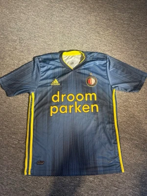 Feyenoord Rotterdam Adidas matchtröja S - Säljer en Feyenoord Rotterdam matchtröja från Adidas i storlek S. Tröjan är marinblå med gula detaljer och har ett prickigt mönster. Klubbmärket är broderat på bröstet och det finns gula ränder längs sidorna. Perfekt för fotbollsfans!