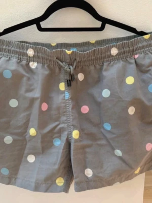 Nikben Grå prickiga shorts med snörning - Säljer ett par grå shorts med färgglada prickar i blått, gult, rosa och vitt. De har elastisk midja med snörning och en bakficka med lock. Perfekta för sommaren och chill dagar vid stranden eller poolen.