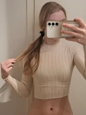 ribbad croppad tröja - Säljer en beige croppad tröja från FB Sister med ribbat mönster och lång ärm. Tröjan har en hög hals med vågig kant och är figurnära i modellen. Perfekt att styla med höga jeans eller kjol.