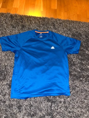 Blå Adidas Climalite t-shirt herr L - Snygg blå t-shirt från Adidas med Climalite-teknologi som håller dig torr och bekväm. T-shirten har en vit Adidas-logga på bröstet och en orange detalj vid halsen. Perfekt för träning eller sportaktiviteter. Den är i storlek L. Den är i bra skick