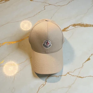 Beige Moncler keps med broderad logga - Snygg beige keps från Moncler med broderad logga framtill. Kepsen har klassisk form med böjd skärm och justerbar baksida. Materialet är slitstark bomull som ger en clean och stilren look. Perfekt för dig som vill ha en enkel men exklusiv accessoar.