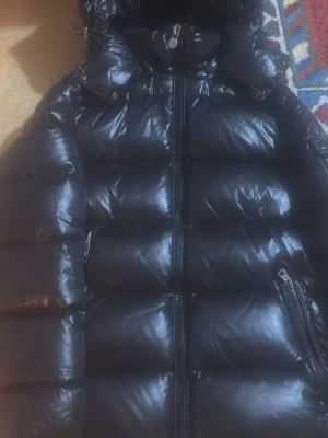 Svart Moncler pufferjacka med huva - Svart pufferjacka från Moncler med glansig yta, huva och dragkedja framtill. Jackan har Moncler-logga på ärmen och praktiska fickor med dragkedja. Perfekt för kalla dagar och har en riktigt schysst streetstil.