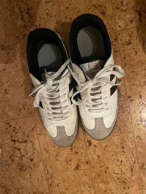 Adidas Samba vita och svarta sneakers - Säljer ett par klassiska Adidas Samba sneakers i vitt med svarta ränder och grå mockadetaljer på tån. Skorna har snörning, platt sula i gummi och rund tå. Insidan är svart och ovandelen är i skinn och mocka. Perfekt för dig som gillar retrostil och sportig vibe.