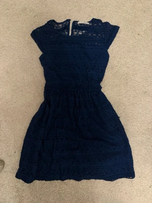 Marinblå spetsklänning från Vila - Säljer en marinblå kortklänning från Vila med vackra spetsdetaljer och rund hals. Klänningen har korta ärmar och markerad midja för en feminin siluett. Perfekt för dig som vill ha en söt och stilren look.
