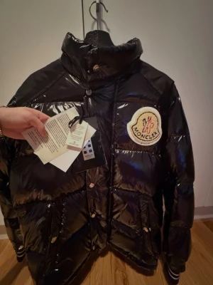 Moncler x Palm Angels jacka - Säljer en svart pufferjacka från Moncler med glansig finish och stor broderad logga på bröstet. Jackan har lila foder, dragkedja och knappar framtill samt höga fickor. Perfekt för dig som vill ha en riktigt snygg vinterjacka med exklusiv känsla.