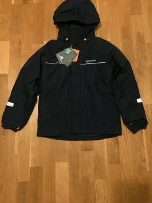 Svart vinterjacka från Didriksons - Säljer en svart vindjacka från Didriksons med huva och orangea detaljer vid knapparna. Jackan har vita reflexdetaljer på ärmar och rygg, samt diskret logga på bröstet. Materialet är polyester och jackan är perfekt för blåsiga dagar. Den är helt ny med prislapp aldrig använd. Extend szize. 1000kr nypris