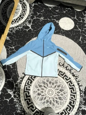 Nike blå nike tech tröja - Nike tech i två nyanser av blått med svart dragkedja och  detalj på ärmen. Jackan har huva, lång ärm och en sportig look. Materialet är polyester och jackan har en normal passform. Perfekt för dig som gillar streetstyle och sport.