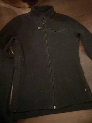 Svart fleecejacka från Texstar XS - Svart fleecejacka från Texstar i storlek XS. Jackan har hel dragkedja framtill, ståkrage och en diskret bröstficka med dragkedja. Materialet är mjukt och värmande fleece, perfekt för kyliga dagar. Snygg och enkel design med långärmade ärmar.