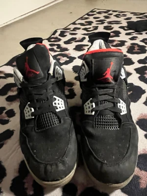 Svarta Nike Jordan 4 sneakers - Nike Air Jordan 4 sneakers i svart med röda detaljer och vit mesh. Skorna har snörning, platt sula och rund tå. Ikonisk Jumpman-logga på plösen och hälen. Materialet är skinn och syntet, med vadderad insida i vitt och rött. Perfekt för dig som gillar streetwear och klassiska sneakers.