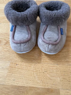 Gråa Shepherd tofflor med ullkant - Mysiga gråa tofflor från Shepherd med mjuk och fluffig ullkant runt öppningen. Utsidan är i mocka och insidan är fodrad med varm ull. Perfekta för kalla dagar hemma och har en enkel, rund tå och platt sula.Ej använda.